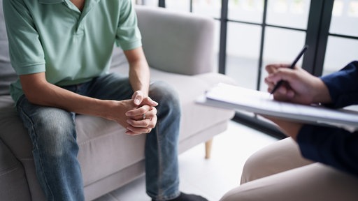 Ein Patient sitzt auf einer Couch, gegenüber sitzt ein Therapeut und macht sich Notizen