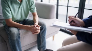 Ein Patient sitzt auf einer Couch, gegenüber sitzt ein Therapeut und macht sich Notizen