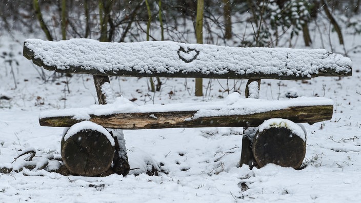 Eine Bank im Wald ist mit Schnee bedeckt. In den Schnee ist ein Herz gezeichnet.