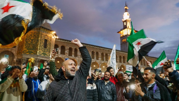 Syrer rufen Slogans und schwenken Fahnen vor der Umayyad-Moschee vor einem Gebet im Vorfeld der Feierlichkeiten zum ersten Jahrestag des Sturzes des Regimes von Assad.