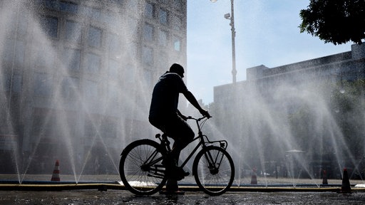 Ein Mann fährt auf einem Fahrrad an sprühenden Wasserschläuchen vorbei, die die Stadt Köln im Rahmen des Hitzeschutz ausgelegt hat.