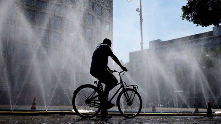Ein Mann fährt auf einem Fahrrad an sprühenden Wasserschläuchen vorbei, die die Stadt Köln im Rahmen des Hitzeschutz ausgelegt hat.