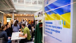 Menschen unterhalten sich bei einer Jobmesse für ukrainische Geflüchtete von der IHK Berlin und der Agentur für Arbeit. Rechts zeigt ein Bildschrin die ukrainische Flagge und den Gruß "Willkommen auf unserer Jobmesse" auf Deutsch und Ukrainisch.