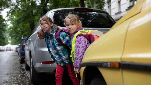 Zwei Mädchen mit Schulrucksack schauen zwischen geparkten Autos hervor
