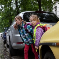 Zwei Mädchen mit Schulrucksack schauen zwischen geparkten Autos hervor