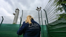 Eine Mitarbeiterin der Ausländerbehörde steht im Außenbereich der hoch umzäunten Außengrenzeinrichtung am Berliner Flughafen unter einem Kamera-Turm.