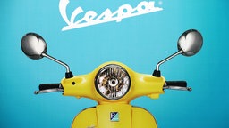 Ausschnitt eines Posters: Eine gelbe Vespa vor einem blauem Hintergrund mit dem typischen Schriftzug "Vespa" in Weiß.