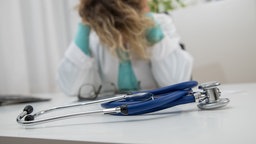 Gestellte Szene: Eine Ärztin sitzt am Tisch und hält ihren Kopf zwischen den Händen. 