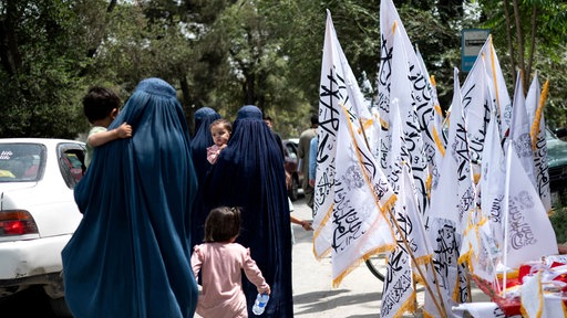 Afghanische Frauen in Burkas gehen an Taliban-Flaggen vorbei. Symbolbild: Vier Jahre nach Machtübernahme der Taliban