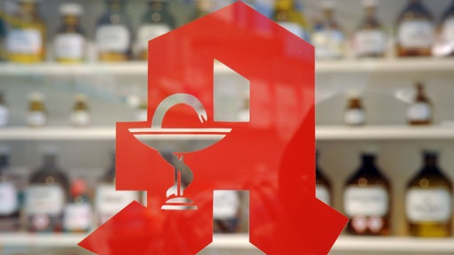 Blick durch eine Scheibe, auf der das rote Apothekenlogo zu sehen ist. Dahinter sind Regale mit Medikamenten zu erkennen.