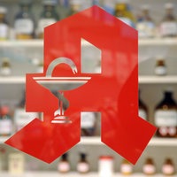 Blick durch eine Scheibe, auf der das rote Apothekenlogo zu sehen ist. Dahinter sind Regale mit Medikamenten zu erkennen.