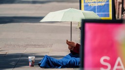 Symbolbild: Ein Mensch sitzt mit Strohut und Schirm in einer Fußgängerzone in der Sonne und bettelt.