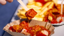 Eine Hand piekt ein Stück Currywurst aus einer klassischen Pappschale auf.