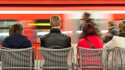 Symbolbild: Menschen sitzen an einem Bahnsteig, an dem eine S-Bahn vorbeifährt.