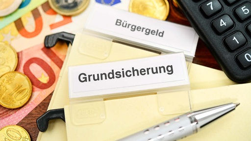Symbolbild: Neben Euroscheinen und Taschenrechner liegen Karten mit der Beschriftung "Bürgergeld" und "Grundsicherung".