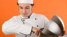 Symbolbild: Ein Mensch mit Kochmütze und Backschüssel vor orangefarbenem Hintergrund (gestellte Szene).