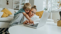 Mutter mit kleinem Kind vor Notebook im Wohnzimmer. 
