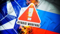 Symbolbild: Ein Warnschild mit der Aufschrift "Hybrid Warfare" zwischen den Fahnen der NATO und Russlands.
