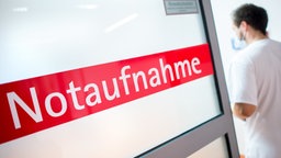 Ein Krankenpfleger steht in einem Krankenhaus neben einer Glastür mit der Aufschrift "Notaufnahme"
