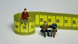 Symbolbild: Spielfiguren an einem Maßband, das Zahlen von 60 bis 70 zeigt.