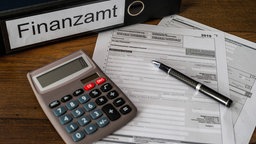 Eine Steuererklärung, ein Taschenrechner und ein Ordner mit der Aufschrift "Finanzamt" liegt auf einem Tisch.