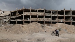 Symbolbild: Ein Mensch schiebt ein Fahrrad an einem zerbombten Gebäude vorbei (25.09.2025, Syrien, Jobar)