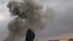 Symbolbild: Rückansicht einer Person, die auf den Rauch übre Teheran nach einem Angriff blickt (02.03.2026).