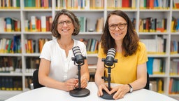 Nicole Bögelein und Gina Rosa Wollinger sitzen an einem Tisch und lächeln in die Kamera. 