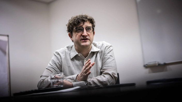 Das Foto von Andreas Stahl zeigt ihn sitzend an einem Tisch: mit kurzem lockigem Haar, dunkler Brille und hellem Hemd.