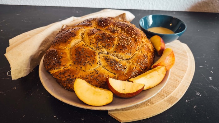 Ein rundes Stück Challah-Brot liegt auf einem Teller, garniert mit Apfelstücken. 