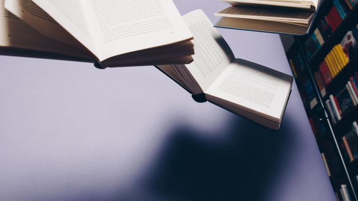 Mehrere aufgeschlagene Bücher schweben über dem Schreibtisch in einer Bibliothek.