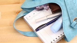 Aus einer blauen Handtasche auf einem Tisch liegend ragen Block, Stift, Lippentift, Deo und Portemonnaie