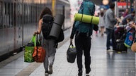 Zwei Menschen mit schwerem Outdoor-Reisegepäck auf den Schultern gehen über den Bahnsteig zu einem Zug.