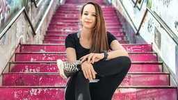 Johanna Pirker, eine junge Frau mit langen Haaren, sitzt auf einer Treppe und hat ein Bein über das andere geschlagen