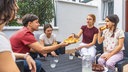 Symbolbild: Mehrere Jugendliche sitzen gemeinsam an einem Tisch im Freien und teilen Pizza.