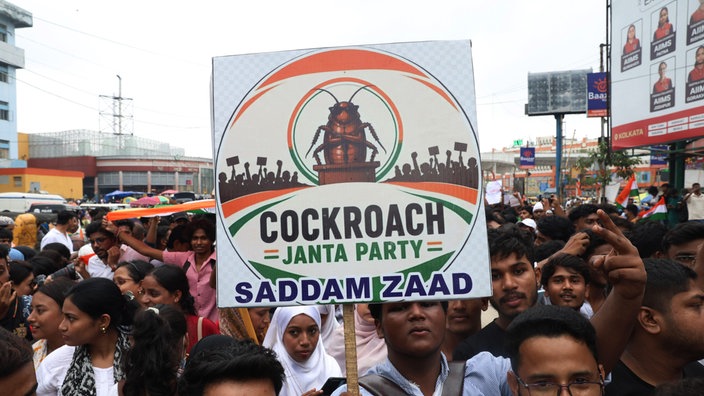 Bei einer Demonstration in Indien hält ein junger Mann ein Schild mit einer Illustration und der Aufschrift "Cockroach Janta Party" in die Höhe.