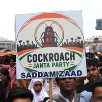Bei einer Demonstration in Indien hält ein junger Mann ein Schild mit einer Illustration und der Aufschrift "Cockroach Janta Party" in die Höhe.