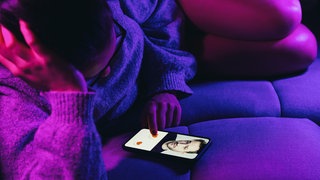 Eine Frau liegt auf einem Sofa; auf ihrem Smartphone ist ihr virtueller Freund zu sehen.
