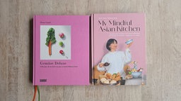 Die beiden rosafarbenen Kochbücher "Gemüse Deluxe" und "My Mindful Asian Kitchen" liegen auf einem gräulichen Holztisch.