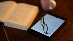 Auf einem Tisch liegt eine Brille auf einem E-Reader neben einem aufgeschlagenen Buch.