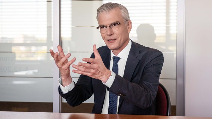 Das Foto zeigt Martin Werding mit gekämmten grauen Haaren, Brille und Anzug mit Krawatte, gestikulierend an einem Tisch sitzend.