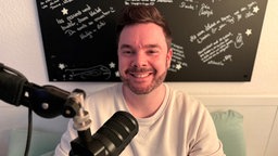 Pascal Hillgruber sitzt aufrecht in seinem Bett vor einem Podcastmikrofon.