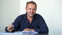 Die Porträtaufnahme von Ralf Südhoff zeigt ihn mit Stift und Notizbuch an einem Schreibtisch.
