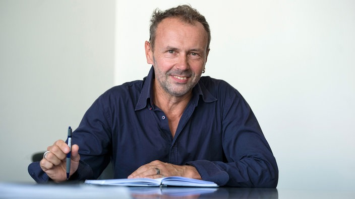Die Porträtaufnahme von Ralf Südhoff zeigt ihn mit Stift und Notizbuch an einem Schreibtisch.