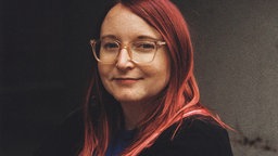 Die Porträtaufnahme zeigt Ramona Wuttig mit roten Haaren, Brille und freundlichem Lächeln.