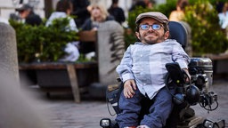 Porträt von Raul Krauthausen, im Hintergrund sitzen Menschen auf einer Terrasse eines Cafés. 