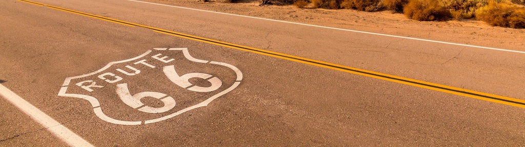 Die legendäre U.S. Route 66 bei Amboy in Kalifornien: Das Route-66-Zeichen auf dem Asphalt, im Hintergrund zieht ein langer Güterzug durch die Mojave-Wüste.
