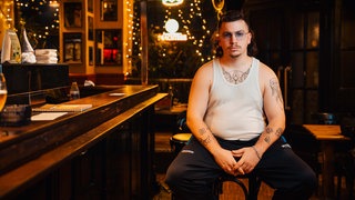 Comedian Ryko sitzt für ein Foto auf einem Hocker in einer Bar. 