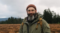Die Porträtaufnahme von Sebastian Berbalk zeigt ihn mit freundlichem Lächeln, schwarz-grauem, langem Vollbart, einer Brille mit dünnem Metallrahmen und einer rostroten Fischermütze, in Outdoor-Kleidung in der Natur.