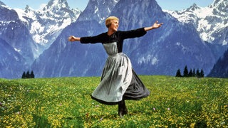 Julie Andrews in einer Szene aus „The Sound of Music“: Eine Frau mit kurzen blonden Haaren im schwarz-weißen Schürzenkleid steht auf einer Alm und hebt die Arme, als würde sie die Natur umarmen.